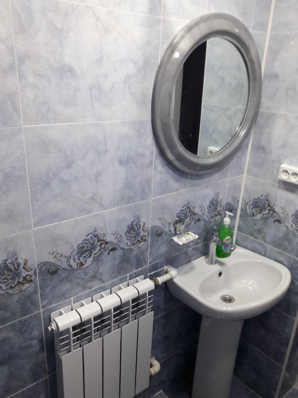 A small bathroom is highlighted by blue tiled walls and floral accents. A round mirror is mounted above a compact sink, with a nearby radiator for heating. Green hand soap is placed on the sink for convenience.