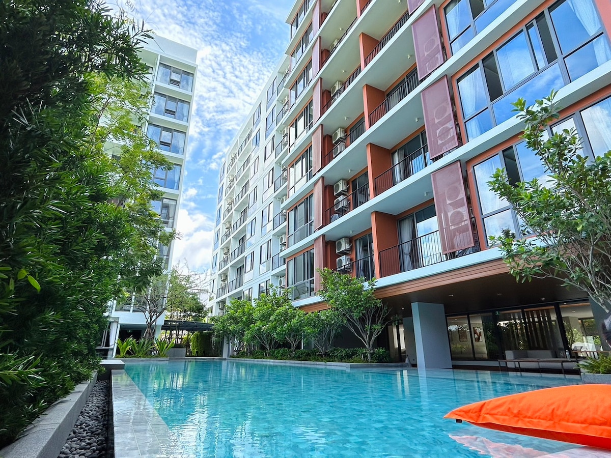 The image displays an outdoor swimming pool surrounded by lush greenery and modern apartment buildings. Comfortable lounge areas are visible, providing a serene space for relaxation. The clear blue water reflects the sky, contributing to a calm and refreshing ambiance within the residential complex.