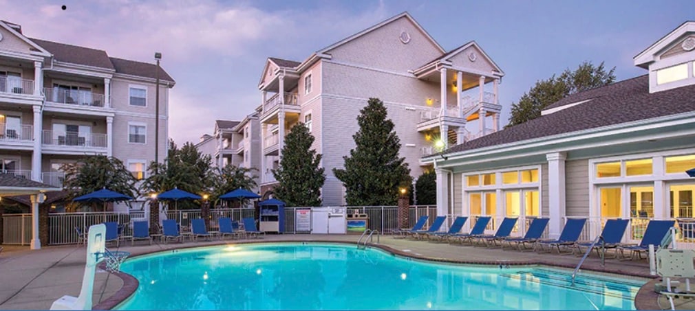 A refreshing swimming pool is surrounded by lounge chairs and umbrellas, creating a relaxed outdoor setting. Nearby, a multi-story building features balconies and lush greenery, contributing to the calm atmosphere. Soft evening light casts a gentle glow over the scene.