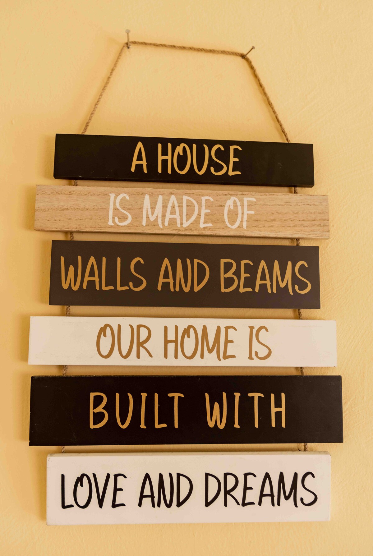 A decorative wall sign, composed of seven wooden planks, features a mix of white and black backgrounds with gold lettering. The phrases express sentiments about home, highlighting the themes of love and dreams. The sign is hung with twine against a warm yellow wall.