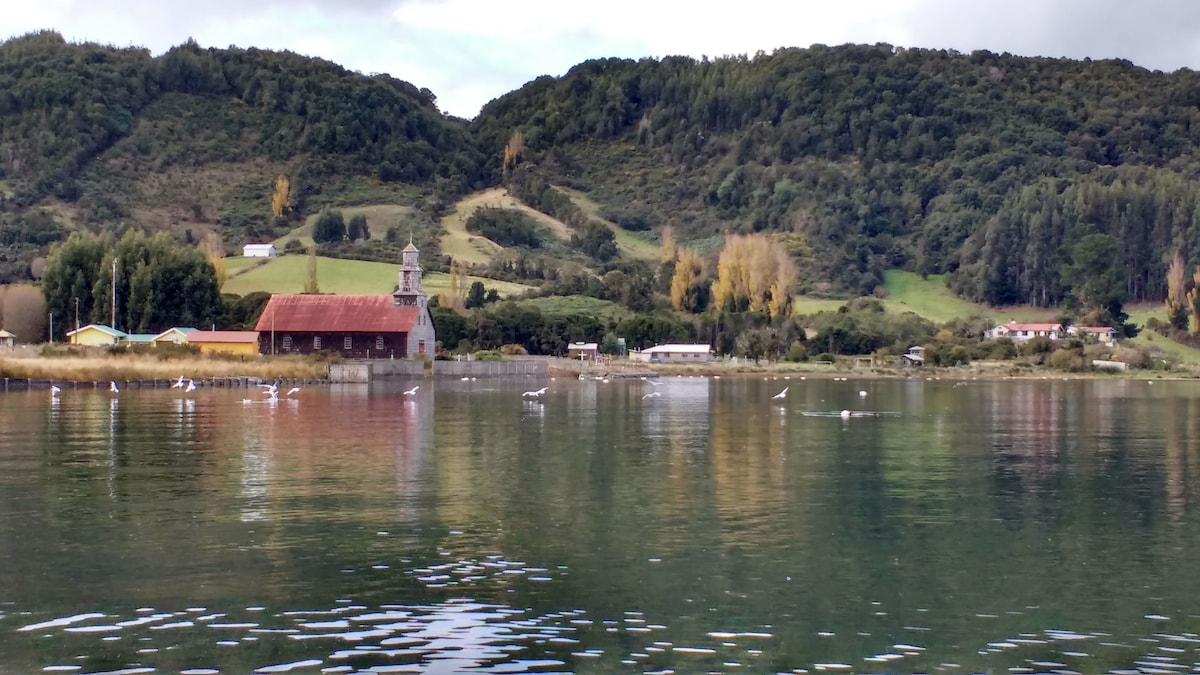 A serene view of the landscape features a calm body of water reflecting the surrounding greenery and a small building with a red roof. Forested hills rise in the background, while several birds can be seen along the water's edge.