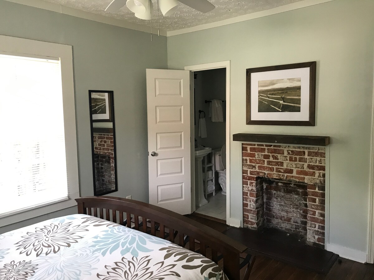 A bedroom features a large window allowing natural light to brighten the space. A cozy bed with a patterned duvet is complemented by a vintage-style exposed brick fireplace. A framed photograph adorns the wall, and a full-length mirror is positioned near the door leading to the bathroom.