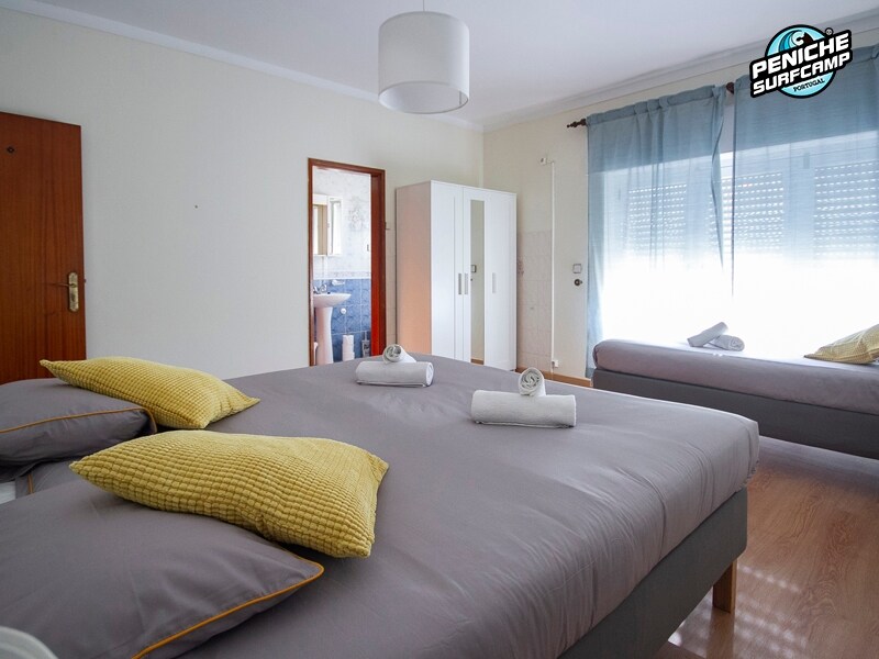 A bright and cozy room contains three individual beds arranged neatly, with two featuring soft yellow accent pillows. Natural light filters through sheer curtains, illuminating the space. A wardrobe stands in the corner, with a door leading to the bathroom visible in the background.