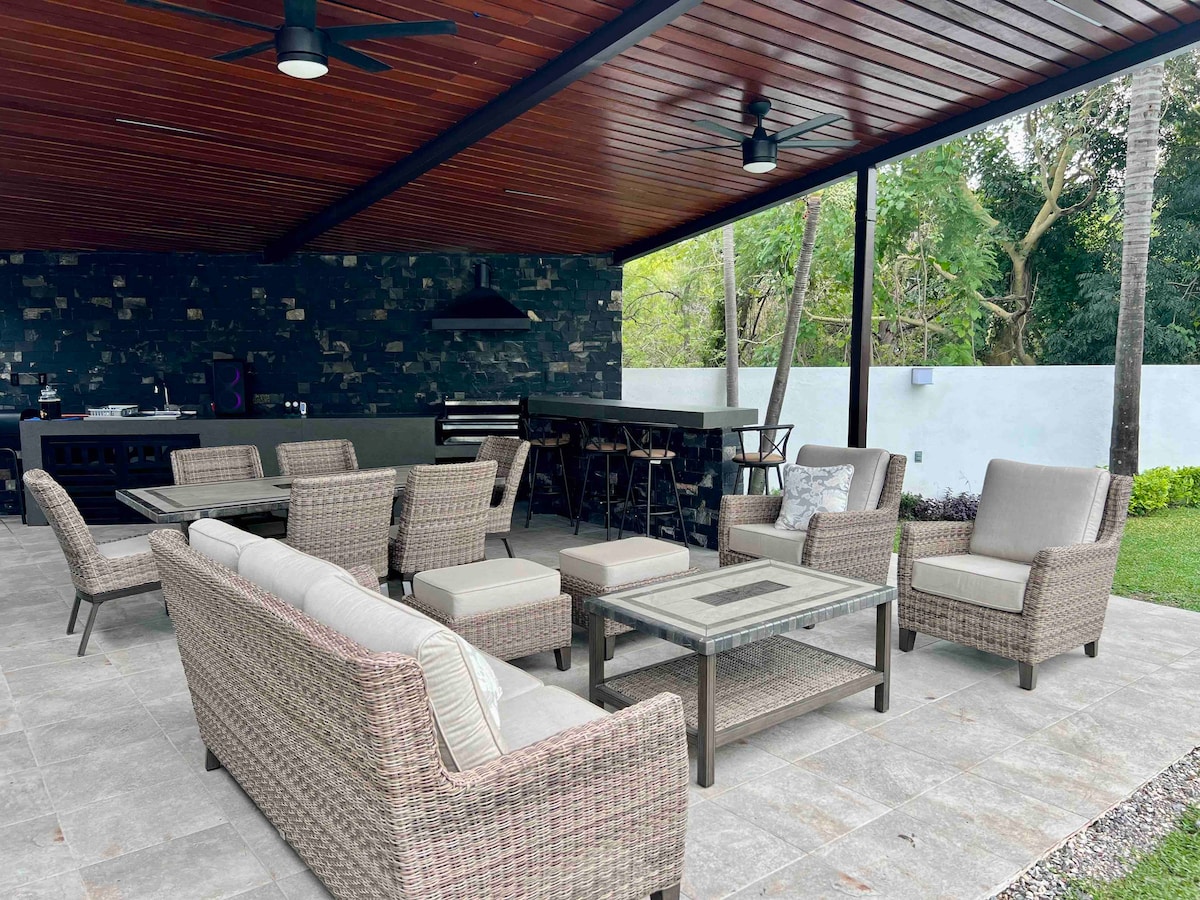 A spacious outdoor area features a covered seating arrangement with multiple wicker chairs and a coffee table. A dining table and bar stools are arranged beneath the sleek wooden ceiling, overlooking a well-maintained garden space in the background.