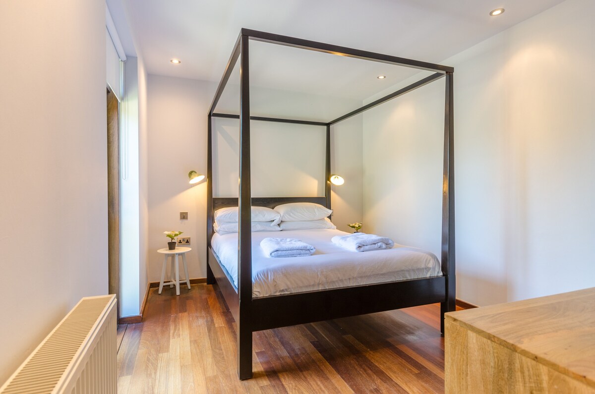 A spacious bedroom features a four-poster bed with crisp white linens and two reading lamps symmetrically placed on either side. Wooden flooring complements the clean, minimalist design, while a small side table adds functionality. Natural light filters in through a nearby window.