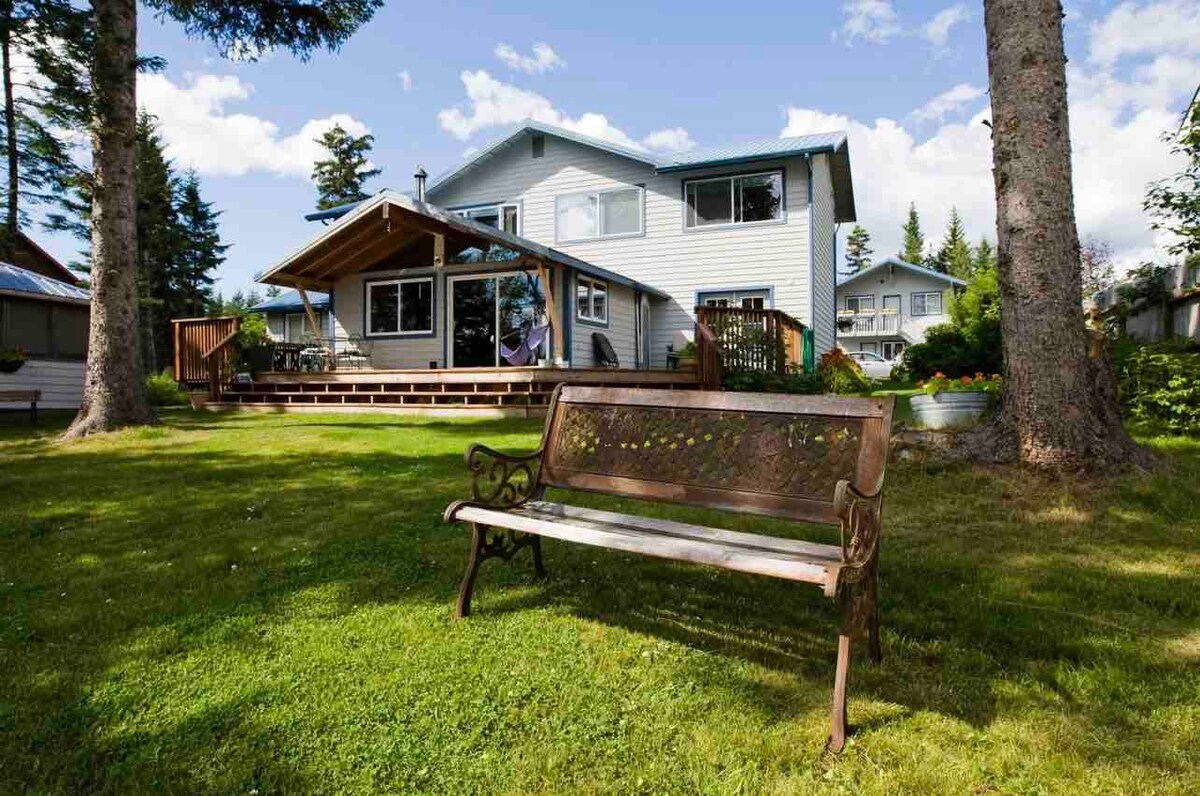 A spacious two-story house is depicted in a tranquil outdoor setting, surrounded by lush greenery. A wooden bench is positioned on the well-maintained lawn, inviting relaxation. The house features a deck with seating areas and large windows, framed by tall trees against a bright blue sky.