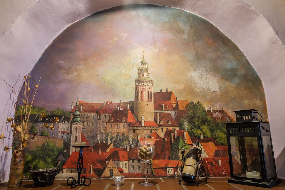 A mural of a historical Czech town is displayed on the wall, featuring a prominent tower amidst a scenic backdrop of rooftops. Various decorative objects, including a lantern and a decorative jar, are arranged in front, adding character to the setting.