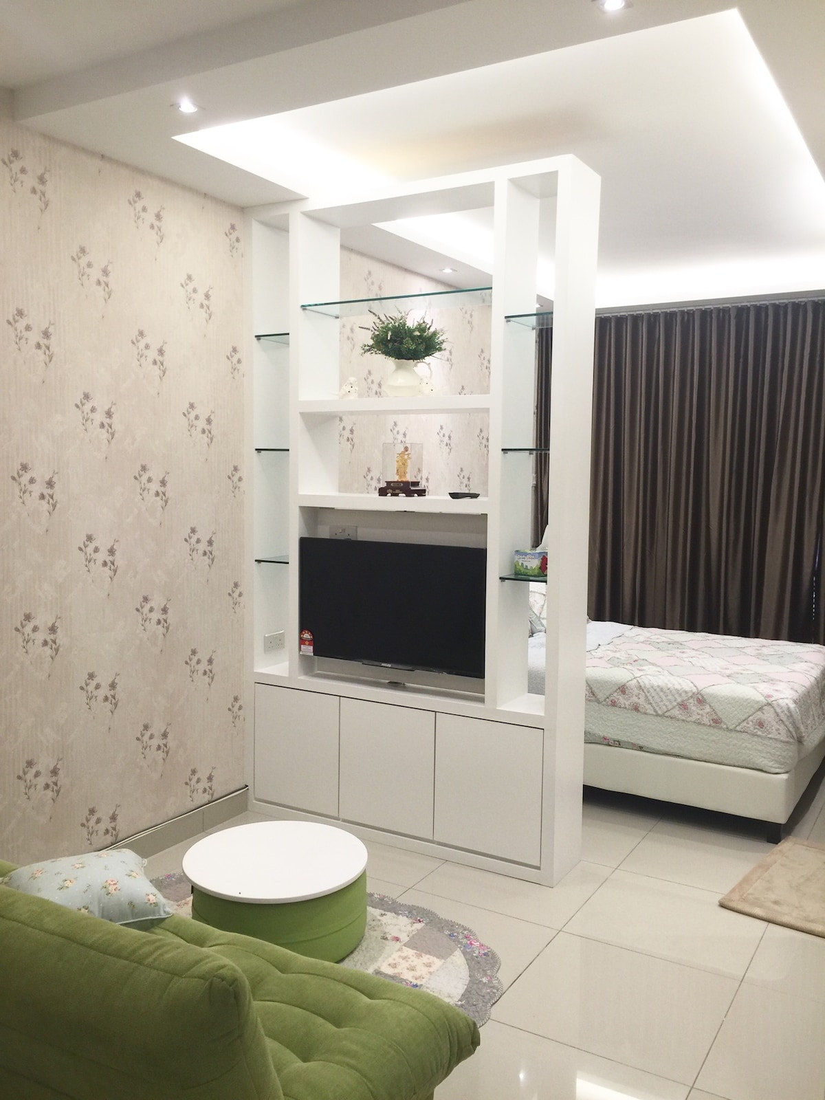 A modern living space is presented with a sleek white shelving unit that holds decorative items and a television. A cozy green chair and circular white table are visible, while a comfortable bed is situated in the background, framed by elegant dark curtains.