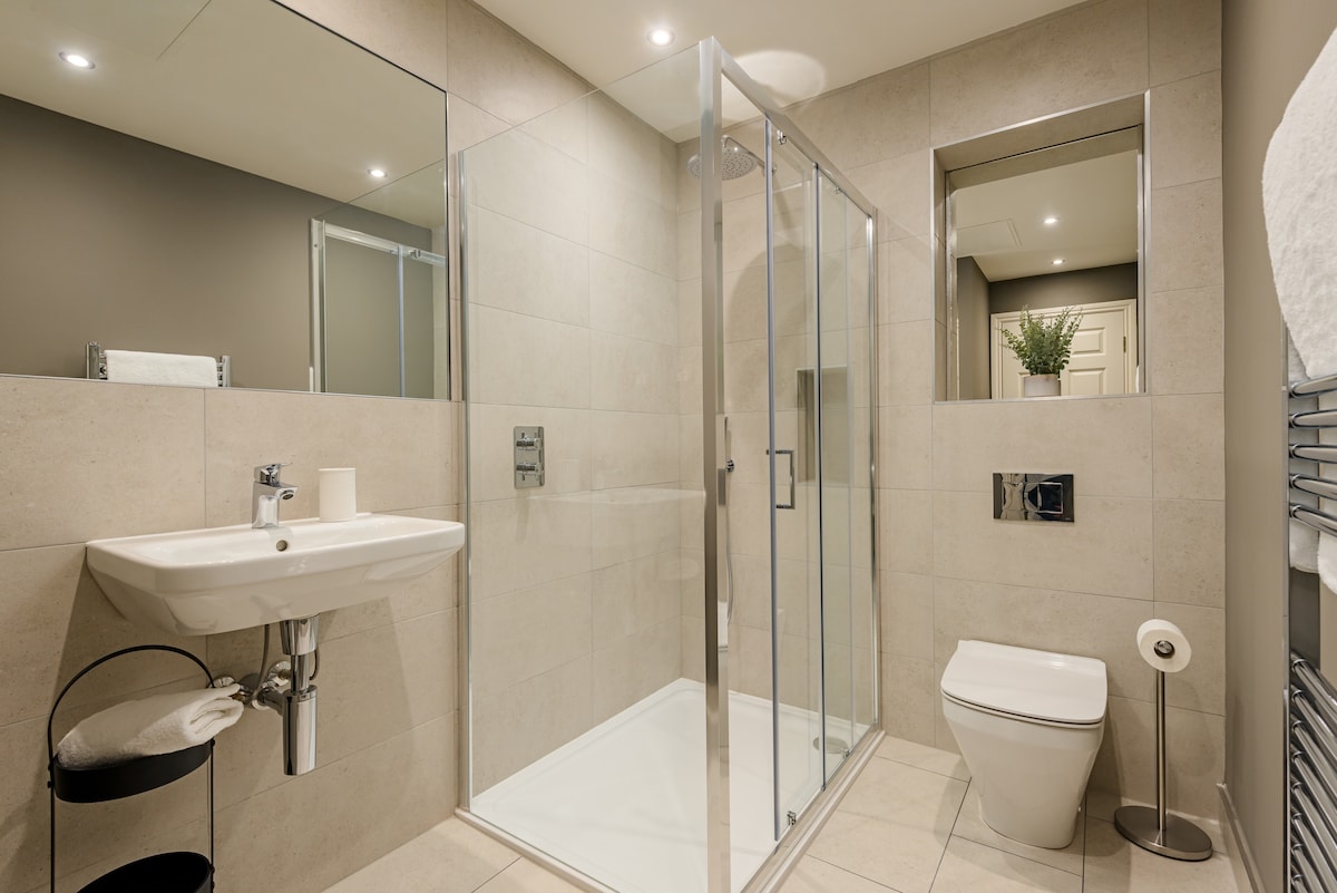 A modern bathroom features a walk-in shower enclosed by glass, complemented by a minimalist sink and a sleek toilet. A heated towel rail is mounted on the wall, with soft towels neatly arranged on a black stand. Natural light is provided through a wall mirror.