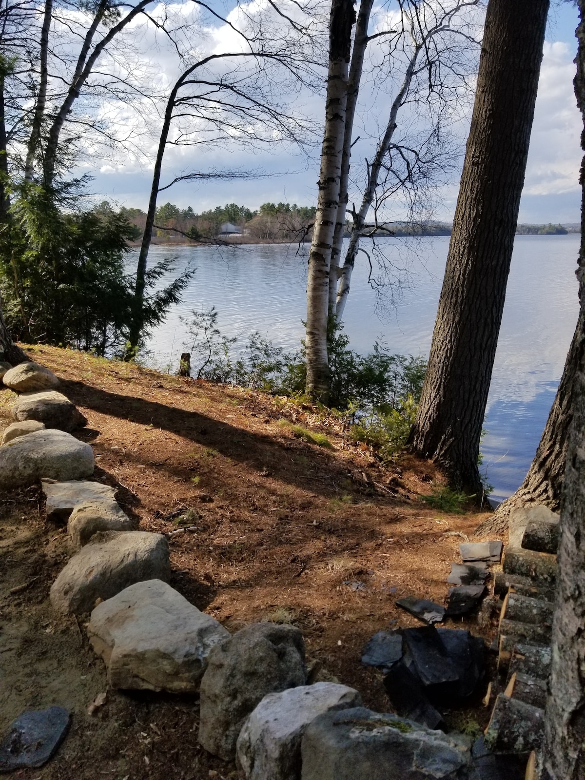A lakeside view is presented with a mix of trees and shrubbery along a dirt path, leading to the water's edge. Smooth stones outline the path, while the lake reflects the sky and surrounding landscape, showcasing a tranquil and natural environment.
