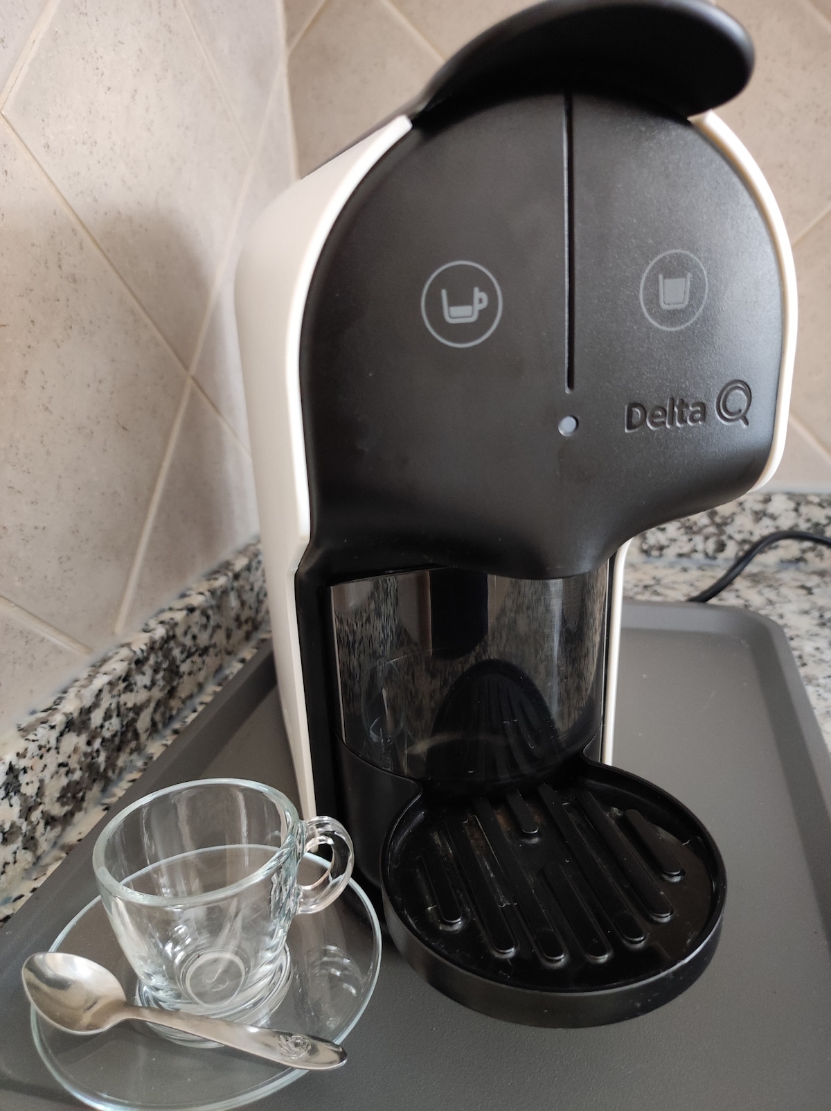 An espresso machine is positioned on a countertop, featuring a sleek, modern design in black and white. A small glass cup and a spoon rest nearby, ready for a fresh brew. The kitchen surface is adorned with a speckled granite pattern.