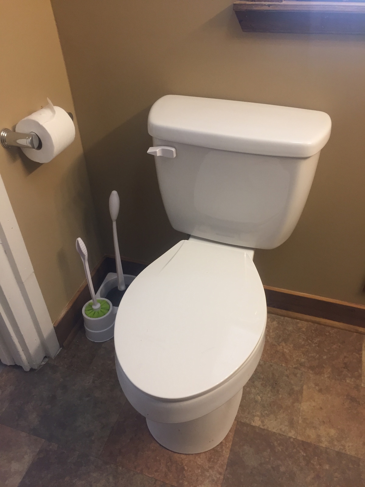 A clean and modern toilet is positioned against a neutral-toned wall. A toilet paper holder is mounted nearby, and two toothbrush holders are placed on the floor beside it. Natural light filters in through a small window, enhancing the space.