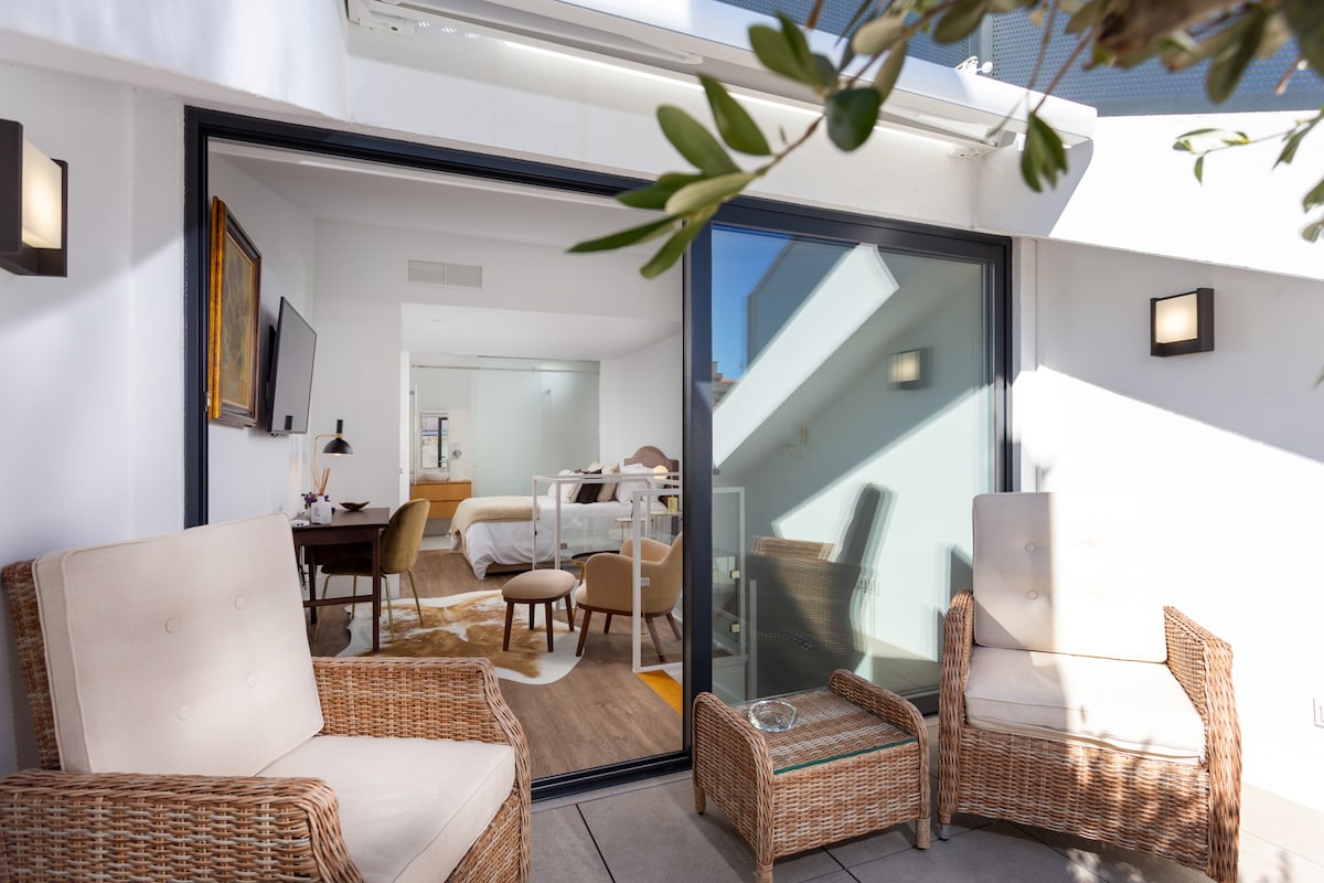 A terrace area features woven armchairs and a small glass-top table, offering a view into the bright interior. Inside, a spacious suite is visible, adorned with modern furnishings and illuminated by natural light from large windows.