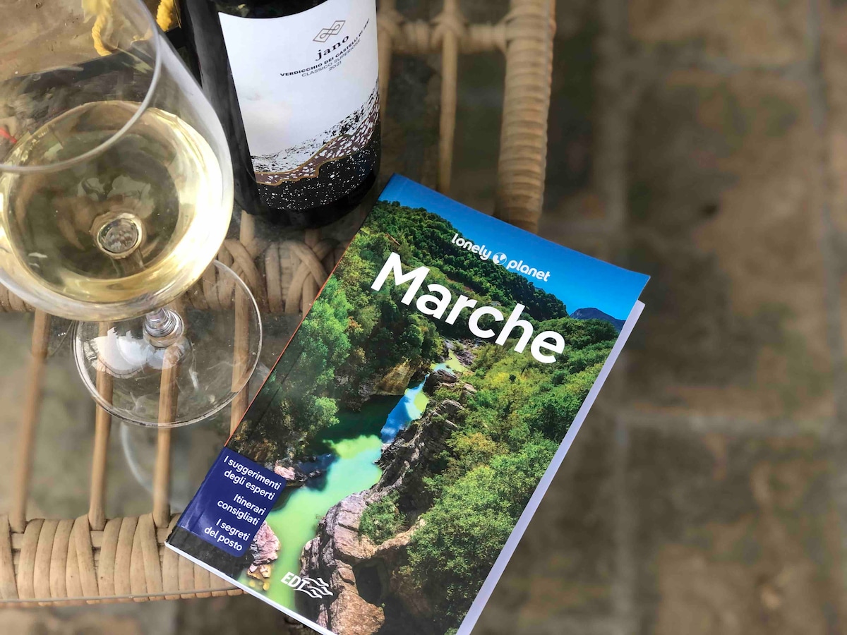 A glass of white wine sits beside a bottle on a woven table. A guidebook titled 'Marche' is open, showcasing scenic landscapes, with vibrant green hills and a flowing river on the cover. The rustic stone surface provides a textured backdrop.