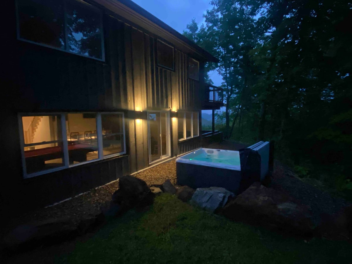 The exterior of the chalet is illuminated in the evening, showcasing a hot tub positioned on a stone patio. Nearby, large windows offer glimpses of the interior, where warm light creates a welcoming atmosphere. The surrounding greenery adds to the property's natural setting.