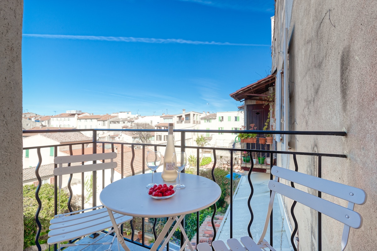 A small balcony features a round table with two white chairs, offering a view of the rooftops and gardens of the Old Town. A glass milk bottle and a plate of red fruit are set on the table, enhancing the inviting atmosphere.