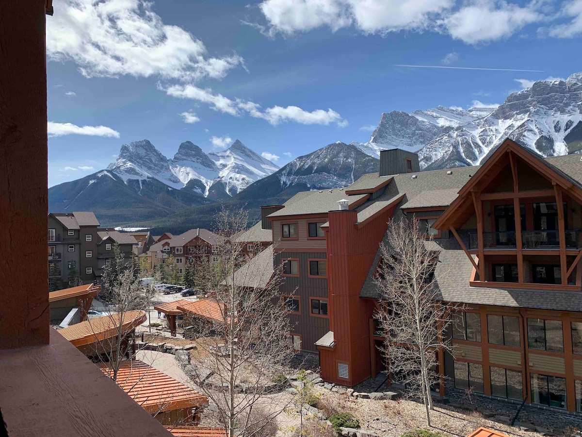 A scenic view showcases the majestic mountains in the background, framed by a clear blue sky accented with a few clouds. The foreground features a blend of architectural structures, highlighting the resort's earthy tones and natural surroundings.