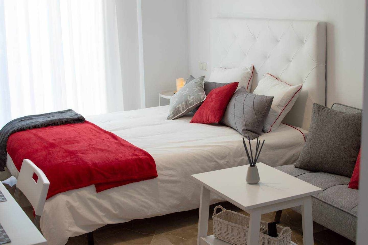 A well-appointed bedroom features a spacious bed adorned with decorative pillows in various hues of red and gray. A cozy gray throw is draped over the bed. A small white table and a gray sofa are positioned nearby, creating a comfortable seating area.