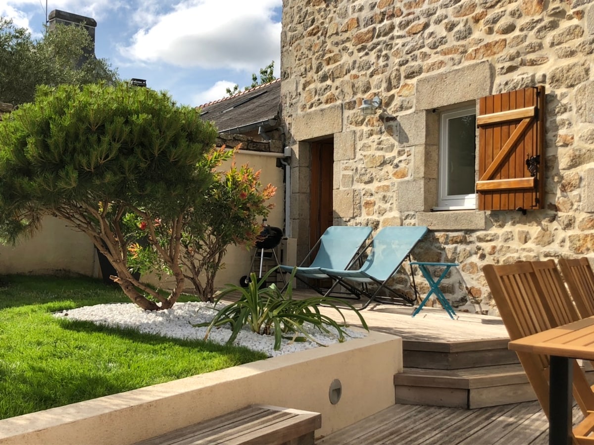 A charming outdoor area features a well-maintained lawn alongside a stone building. Two blue lounge chairs are positioned on a wooden deck, with a small table nearby. Bushes and greenery are present, adding natural elements to the space. Sunlight illuminates the scene.
