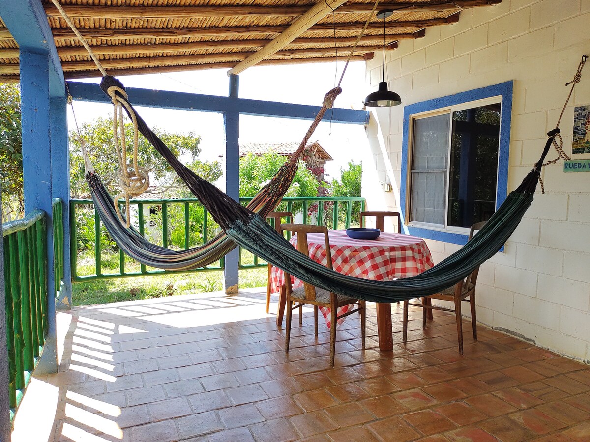 A covered outdoor area features two hammocks gently swaying on either side of a wooden table set with a checkered tablecloth. Green railings border the space, and a light fixture hangs above, with views of lush greenery visible in the background.