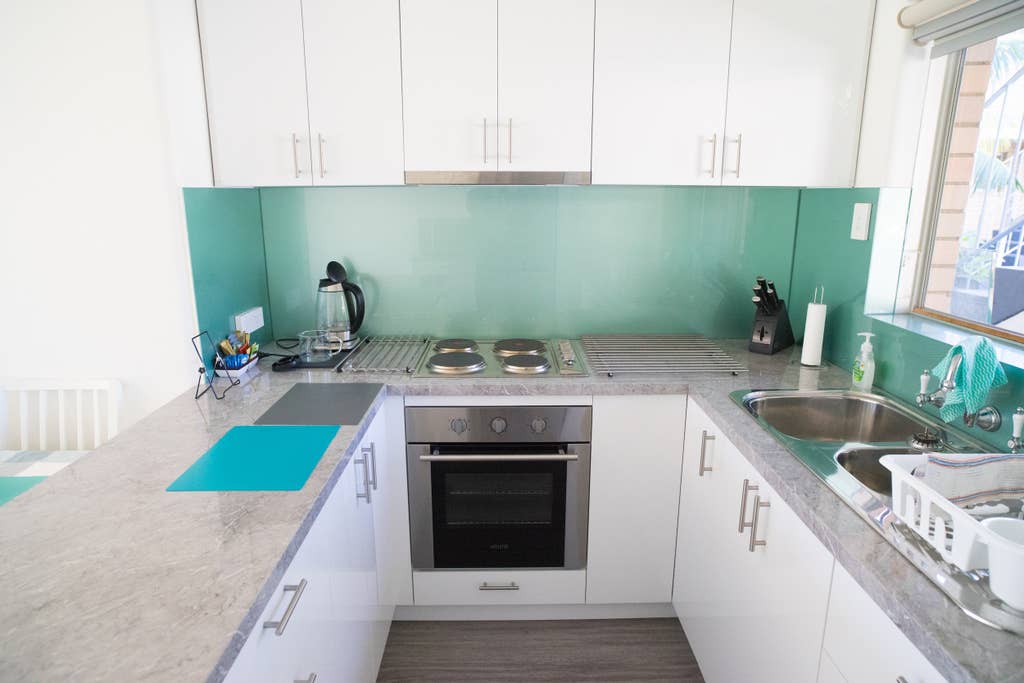 The kitchen features modern cabinetry and a sleek countertop. Stainless steel appliances, including an oven and stovetop, are visible. A kettle and kitchen utensils are neatly arranged, with a window allowing natural light to illuminate the space.