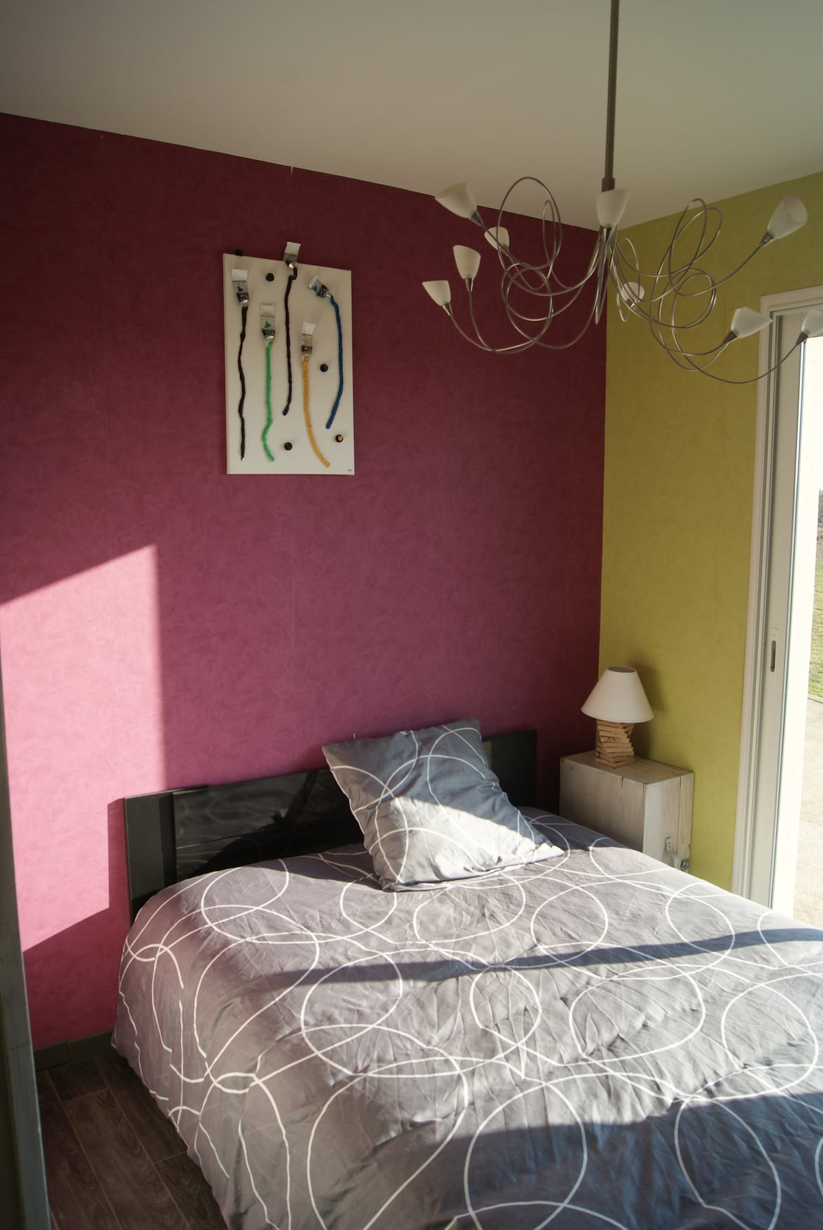 A cozy sleeping area is depicted with a spacious bed dressed in light gray linens featuring circular patterns. One wall is painted in bold pink, while another is in soft green. A small bedside table and a lamp are visible, along with a modern light fixture.