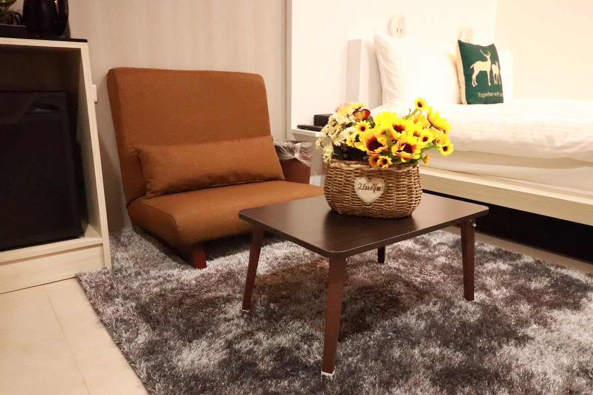 A simple, cozy seating area features a brown chair positioned next to a small table. A wicker basket filled with vibrant sunflowers rests on the table, complementing the soft gray rug and the light-colored bedding visible in the background.
