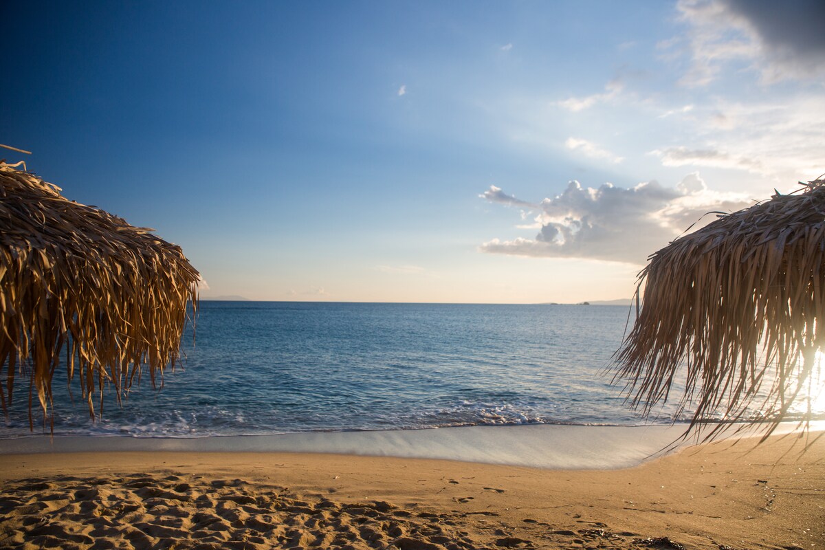 The image captures a serene beach scene featuring two thatched umbrellas framing a calm sea. Gentle waves lap against the golden sand, while a clear sky with soft clouds reflects the warm hues of sunset. The peaceful atmosphere signals a quiet retreat by the water.