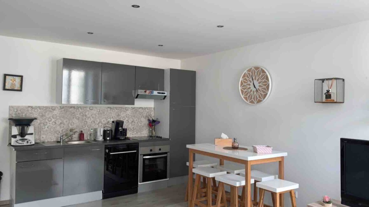 A modern kitchen area features sleek gray cabinetry and a spacious countertop. Appliances include a refrigerator, stovetop, and coffee maker. A small dining table with four wooden stools is positioned nearby, complemented by decorative wall art and ambient lighting.