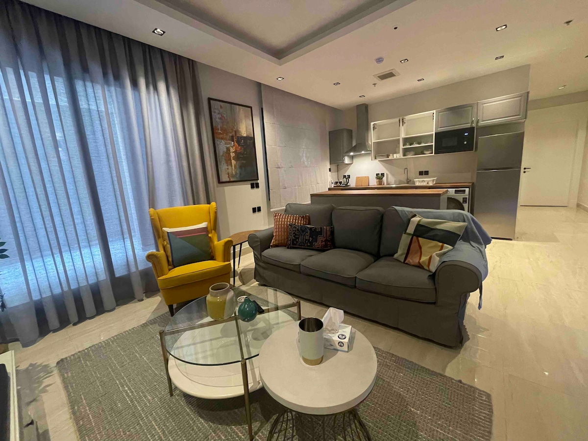 The living area includes a modern gray sofa with colorful cushions and a vibrant yellow armchair. A glass coffee table is positioned in front, while a kitchenette is visible in the background. Natural light filters through sheer curtains, enhancing the open ambiance.