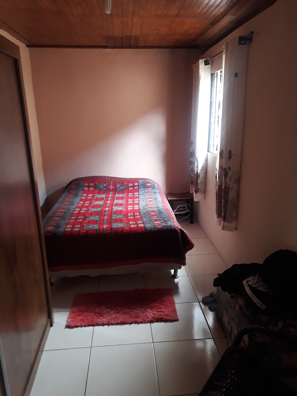 A simple bedroom displays a spacious bed covered with a red patterned blanket. Natural light enters through a window adorned with patterned drapes. The floor is tiled, and clothing is seen resting on a nearby chair, contributing to the room's laid-back feel.