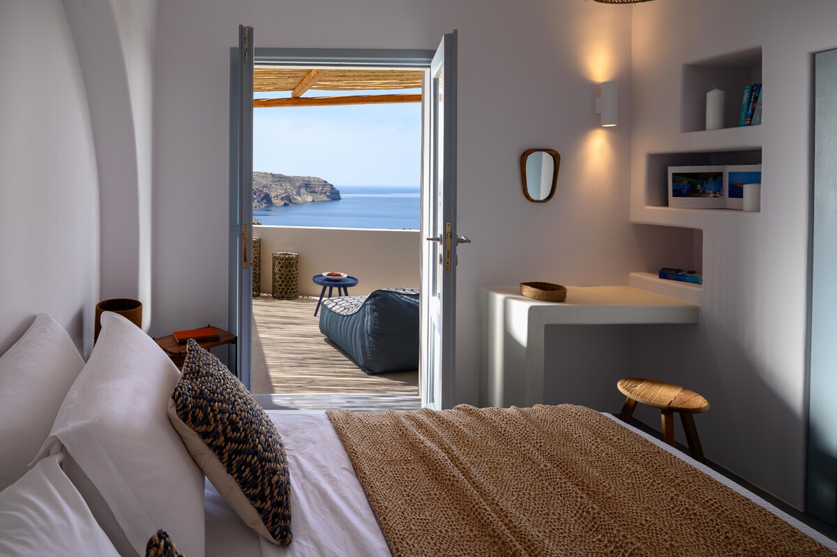 A serene bedroom is viewed from the interior, featuring a spacious bed with neutral linens. A doorway opens to a private terrace with coastal views. Simple furniture includes a stool and a small table. Soft, natural light fills the room, highlighting the clean lines and modern decor.