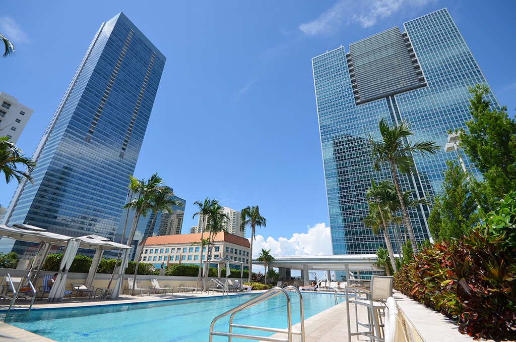 A serene swimming pool is surrounded by lush greenery and modern high-rise buildings. Tall palm trees provide shade next to the pool area, which features lounge chairs and shaded cabanas. The bright blue sky enhances the inviting ambiance of this urban oasis.