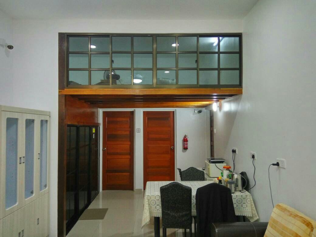 The common living area is highlighted by a light-coloured table with patterned linens, surrounded by four dark chairs. A kitchenette is visible in the background, featuring countertop appliances. Two wooden doors lead to private spaces. An upper loft area is observed through large windows.