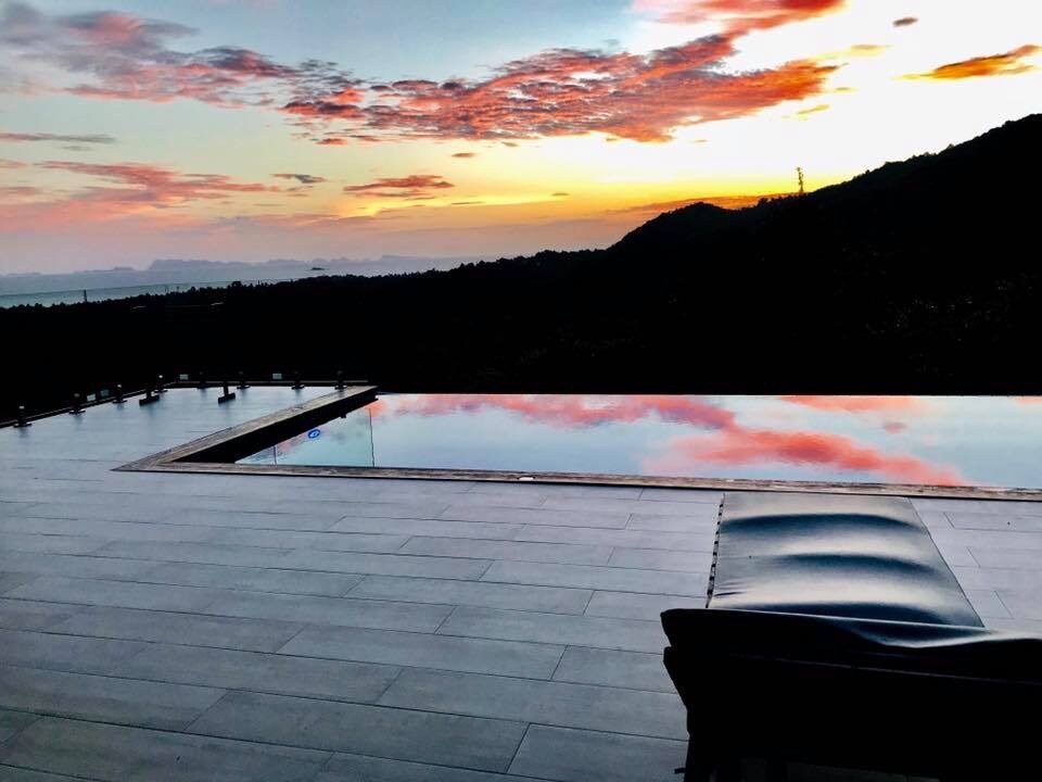 An infinity pool reflects the vibrant colors of a sunset, blending shades of orange and pink across the water's surface. A lone lounge chair is positioned nearby on the large tiled patio, with hills and a distant ocean visible in the background.