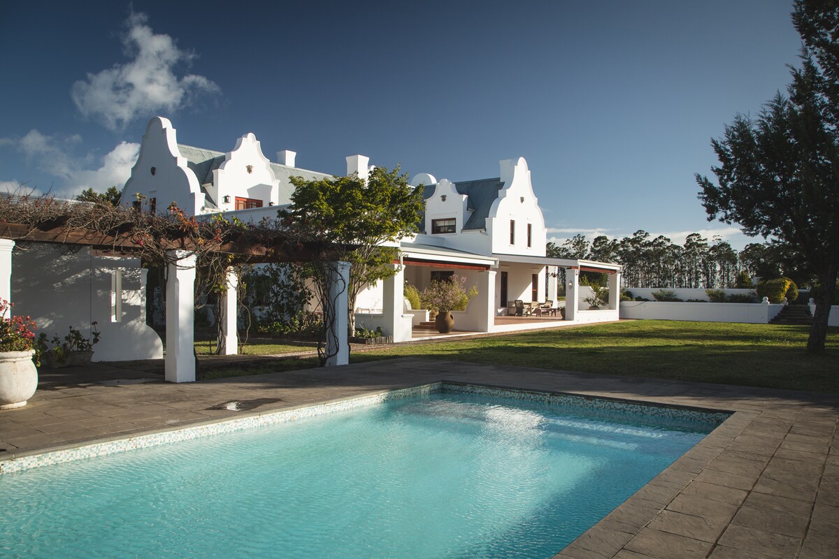 The exterior of a spacious Cape Dutch villa is shown, featuring distinctive gable rooftops and a covered patio. A swimming pool is positioned in the foreground, reflecting the surrounding landscape. Lush lawns and trees create a serene environment, inviting outdoor relaxation.
