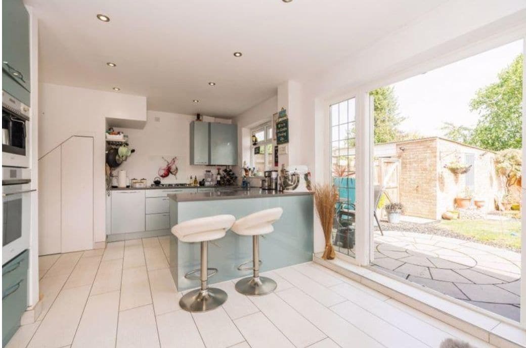 A modern kitchen is presented, featuring light blue cabinetry and sleek stainless-steel appliances. Two bar stools are situated at the kitchen island. Large sliding glass doors offer views of the outdoor space, allowing natural light to fill the area.