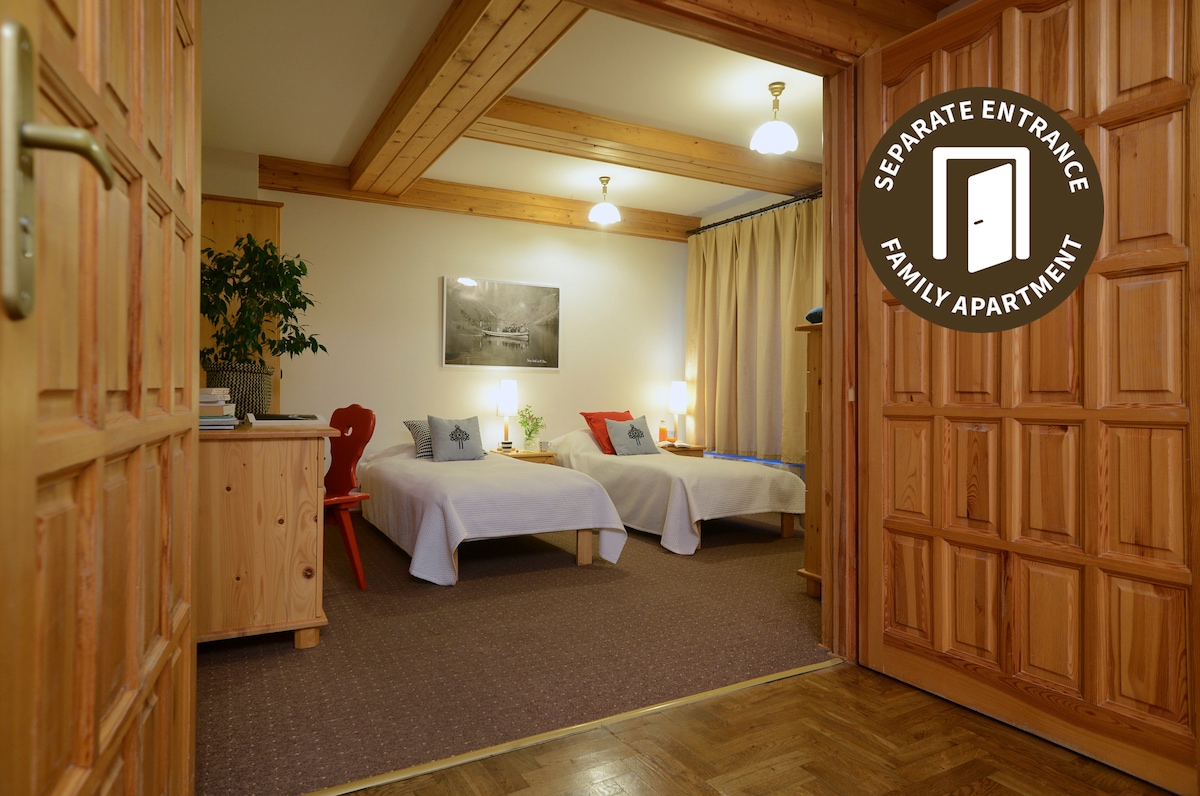 An entrance opens to a spacious family apartment featuring two beds and minimalistic decor. Soft lighting illuminates the room, complemented by light wood accents and a large artwork on the wall. A small table and plant add a touch of warmth to the space.