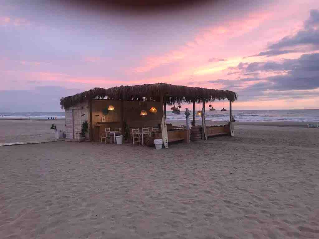 A beachside structure with a thatched roof stands on sandy shores, illuminated by hanging lights. Soft pastel hues of sunset create a serene backdrop, while the gentle waves can be seen lapping at the shoreline in the distance.