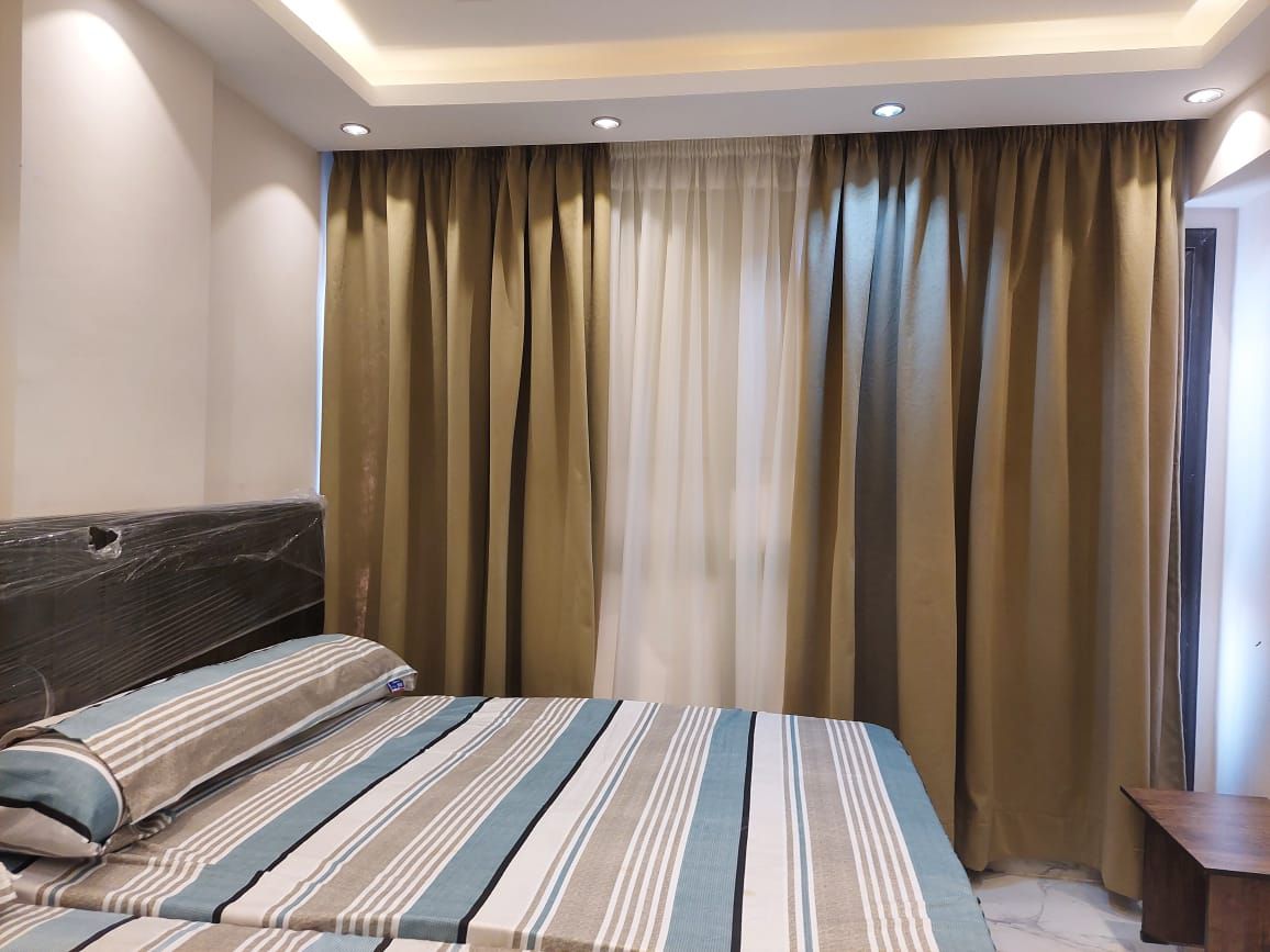 A well-lit bedroom features a spacious bed with a striped duvet. Soft, neutral curtains frame a window, allowing natural light to enter the room. The walls are accented with subtle lighting, adding a calming ambiance to the space.