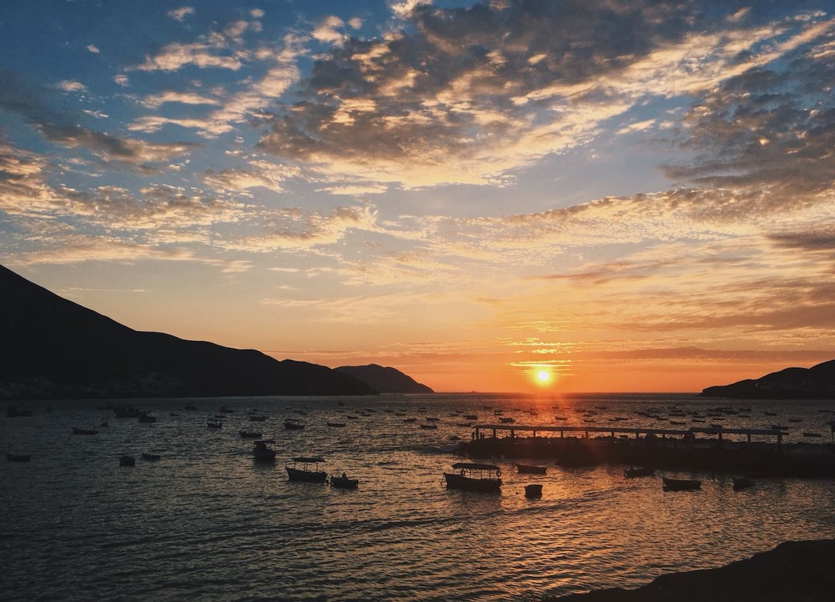 A serene seascape captures a vibrant sunset over calm waters, where numerous small boats are anchored. The sky is filled with scattered clouds, beautifully reflecting the warm hues of the setting sun, while distant hills frame the landscape.