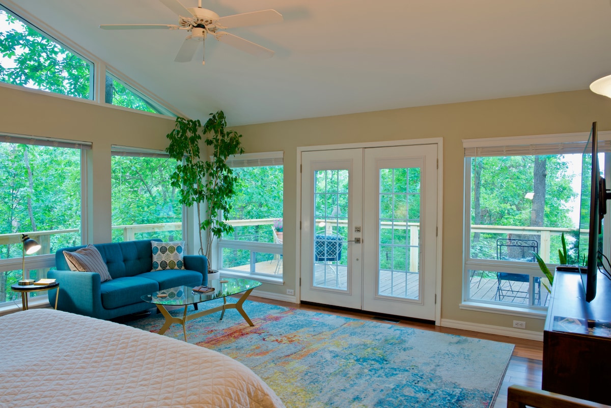A bright and airy space is filled with natural light from multiple windows, showcasing a view of lush greenery. A comfortable sofa sits next to a coffee table on a colorful area rug, with glass doors leading to a private deck.