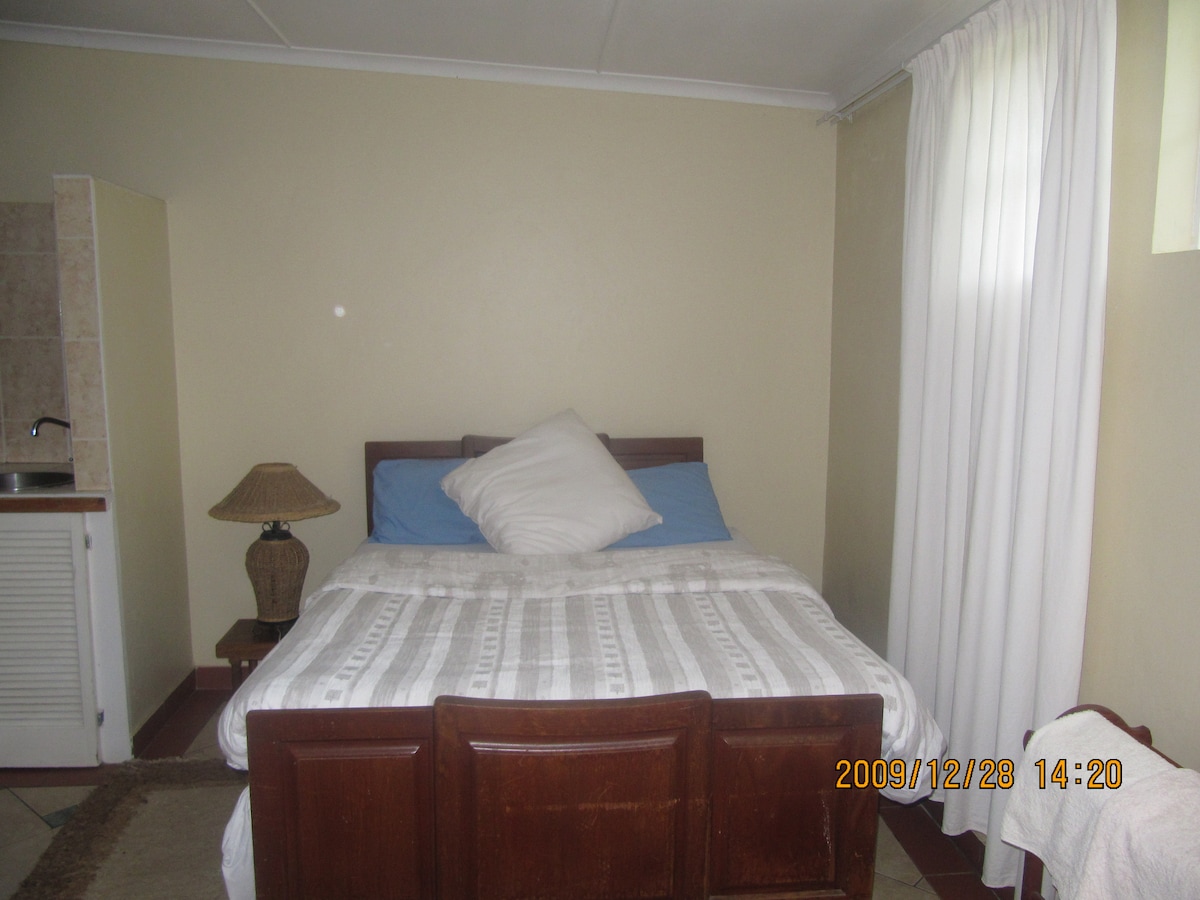 A cozy room features a comfortable bed covered with a light blanket and a decorative pillow. Natural light enters through a window with sheer white curtains. A small kitchenette with a sink is visible, and a bedside lamp adds a warm touch.