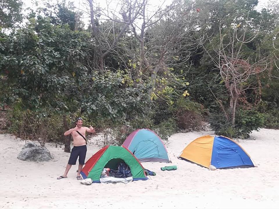 Two colorfully pitched tents are set up on a sandy beach, surrounded by lush greenery. A person is seen adjusting their stance near the tents, enjoying the outdoor setting. The atmosphere suggests a relaxed camping experience in a natural environment.