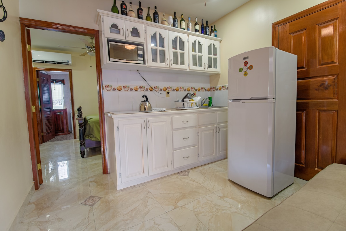 A kitchen area features white cabinetry with glass-paneled doors, a stainless steel refrigerator, and an electric stove. Counter space is utilized for preparing meals, while both a kettle and utensils are visible. The glossy tiled floor reflects the ambient light, enhancing the space's openness.
