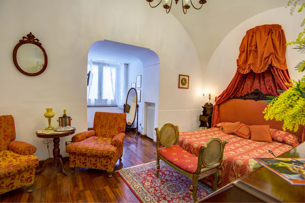 The room features a spacious layout with a decorative canopy bed, complemented by vintage furniture including two armchairs and a small table. Natural light filters through a window, highlighting the hardwood floor and charming decor. A mirror and wall art enhance the inviting atmosphere.