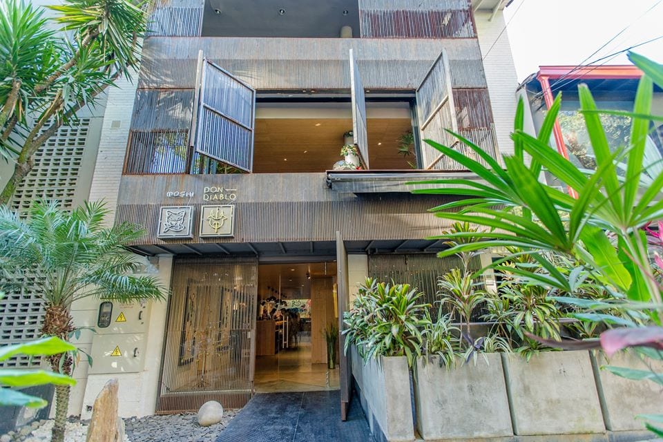 A modern building entrance features large wooden doors flanked by lush green plants. The open doors reveal a welcoming interior, with decorative elements visible inside. A stone pathway leads from the entrance, surrounded by a landscaped area.