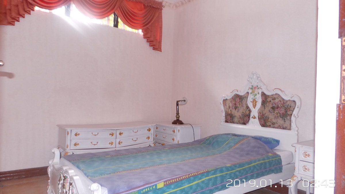 A simple bedroom features light-colored walls with a soft floral accent on the headboard. A spacious bed with a blue and purple coverlet is positioned near a window, allowing natural light to filter in. A white dresser complements the decor, providing additional storage.