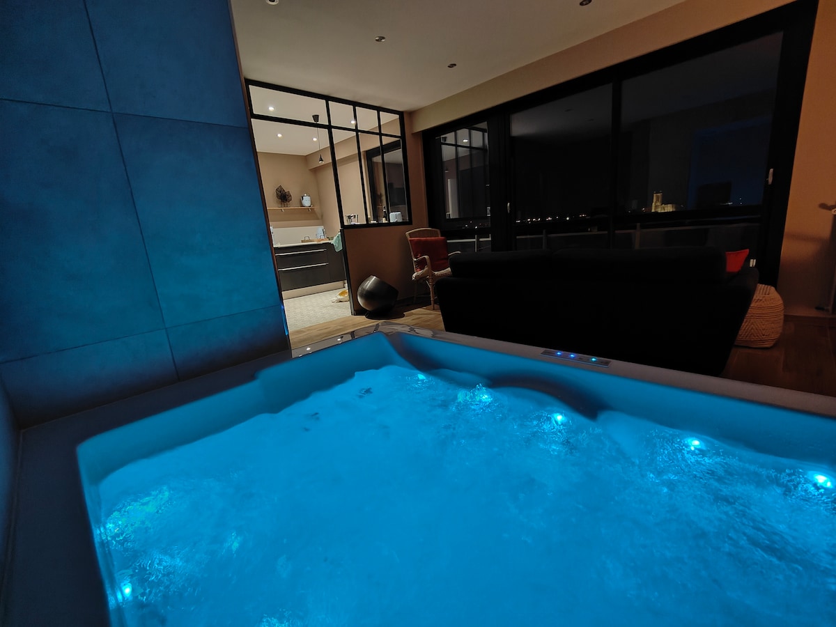 A spacious hot tub is featured, illuminated with soft blue lighting. The view extends to a well-lit open area with large glass doors, revealing a modern kitchen and seating area, creating an inviting ambiance.