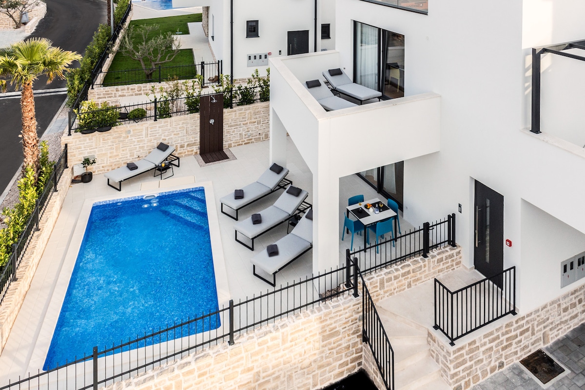 An outdoor area is showcased, featuring a clear blue swimming pool surrounded by several loungers. A patio table and chairs are positioned nearby, offering a space for relaxation. Lush greenery and well-maintained landscaping add to the serene setting.